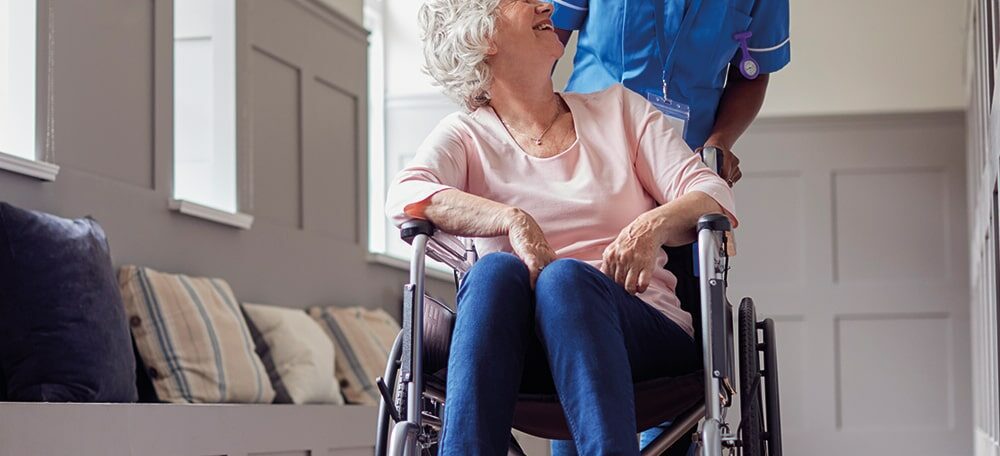 Female care worker in uniform pushes a senior woman in a wheelchair.